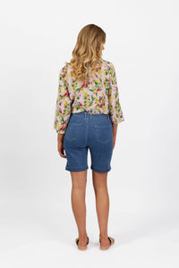 A woman with wavy blonde hair stands facing away, wearing a floral blouse and Vassalli Havana Short - Blue Hue Denim, paired with sandals, against a plain white background.