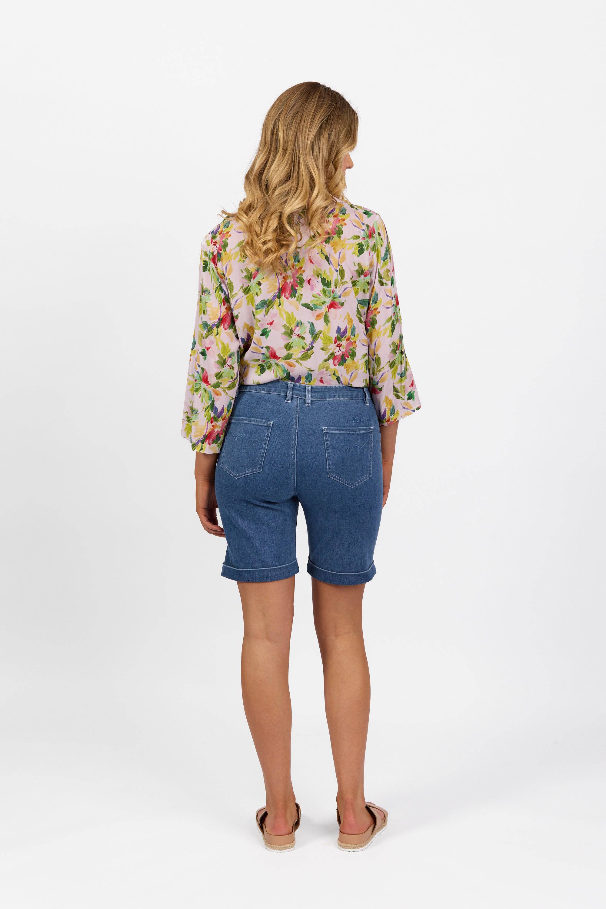 A woman with wavy blonde hair stands facing away, wearing a floral blouse and Vassalli Havana Short - Blue Hue Denim, paired with sandals, against a plain white background.