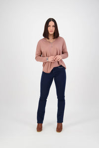 A woman with straight dark hair stands facing forward, wearing a light pink long-sleeve top, Vassalli Urban Jean in mid blue, brown ankle boots, and a necklace, against a plain white background.