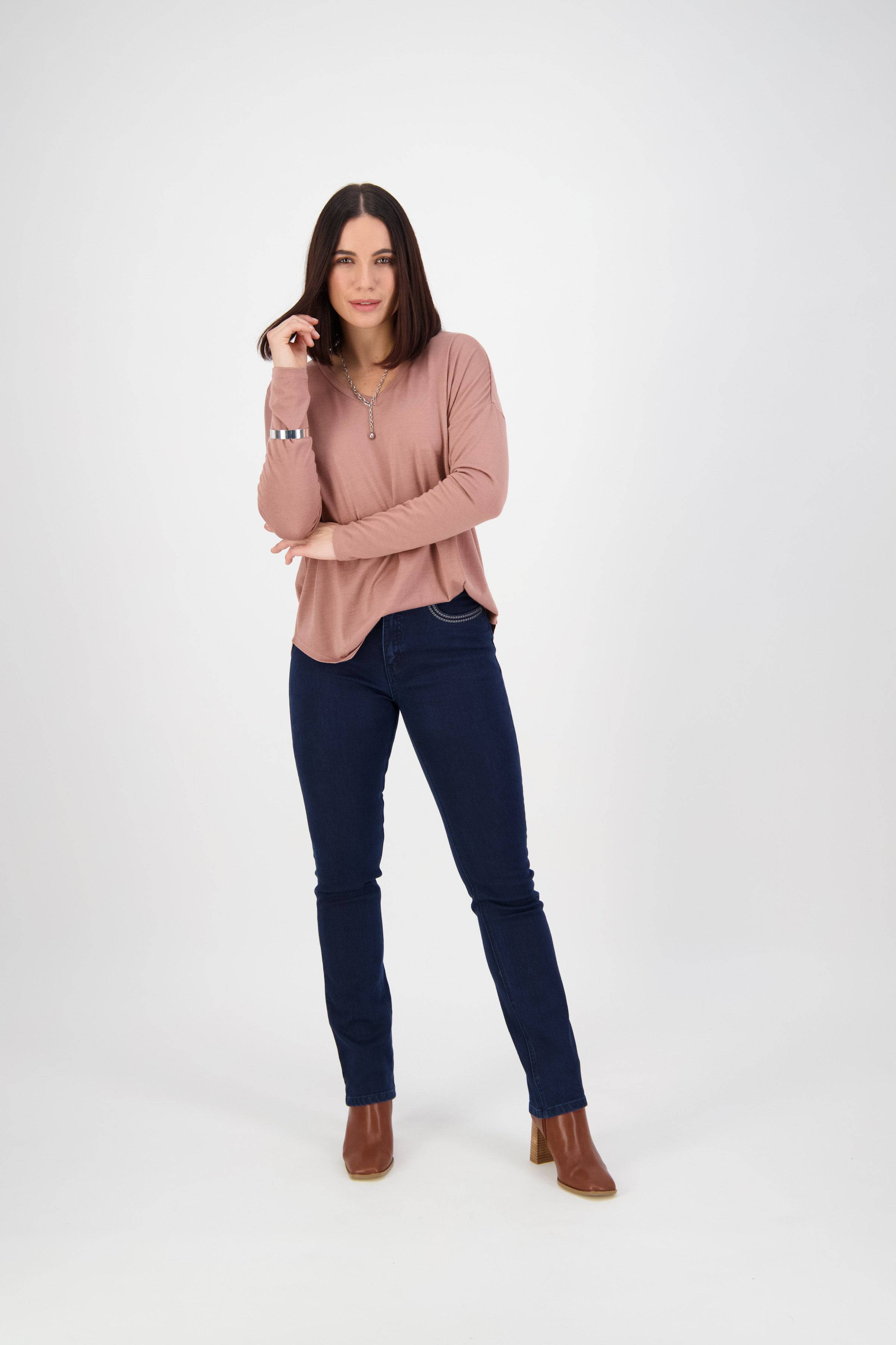 A woman with straight dark hair smiles in a light pink long-sleeve top, Vassalli Urban Jean - Mid Blue, and brown ankle boots against a plain white background, with one hand touching her hair.