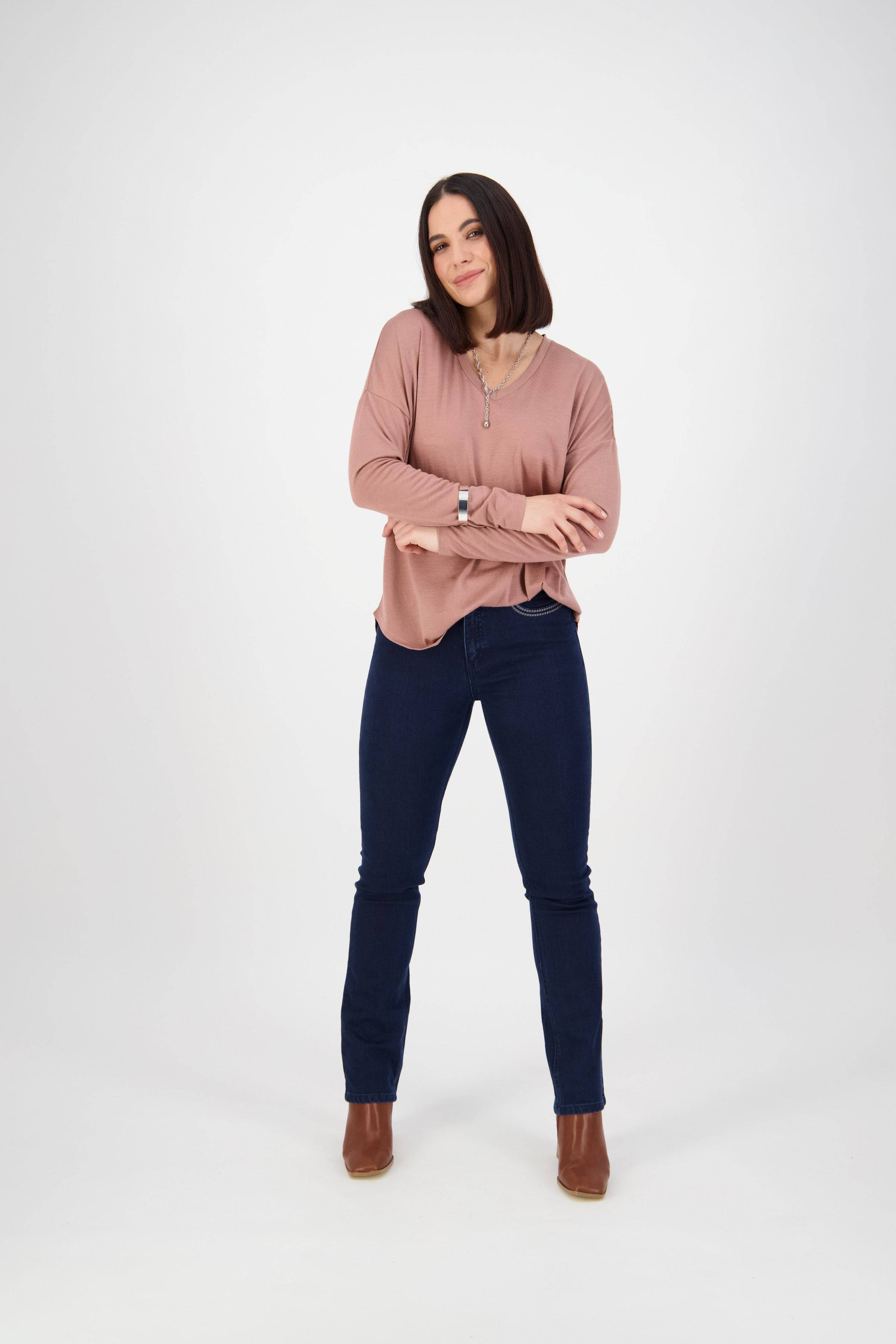 A woman with straight dark hair wears a light pink long-sleeve top, Vassalli Urban Jean in Mid Blue, and brown ankle boots, standing confidently with arms loosely folded in a studio with a white background.