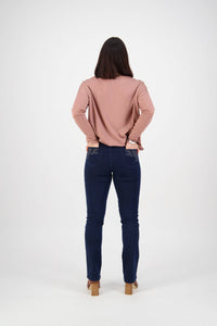 A woman with straight dark hair stands facing away, hands on hips. She wears a long-sleeved pink top, Vassalli Urban Jean - Mid Blue, and tan heeled shoes against a plain white background.