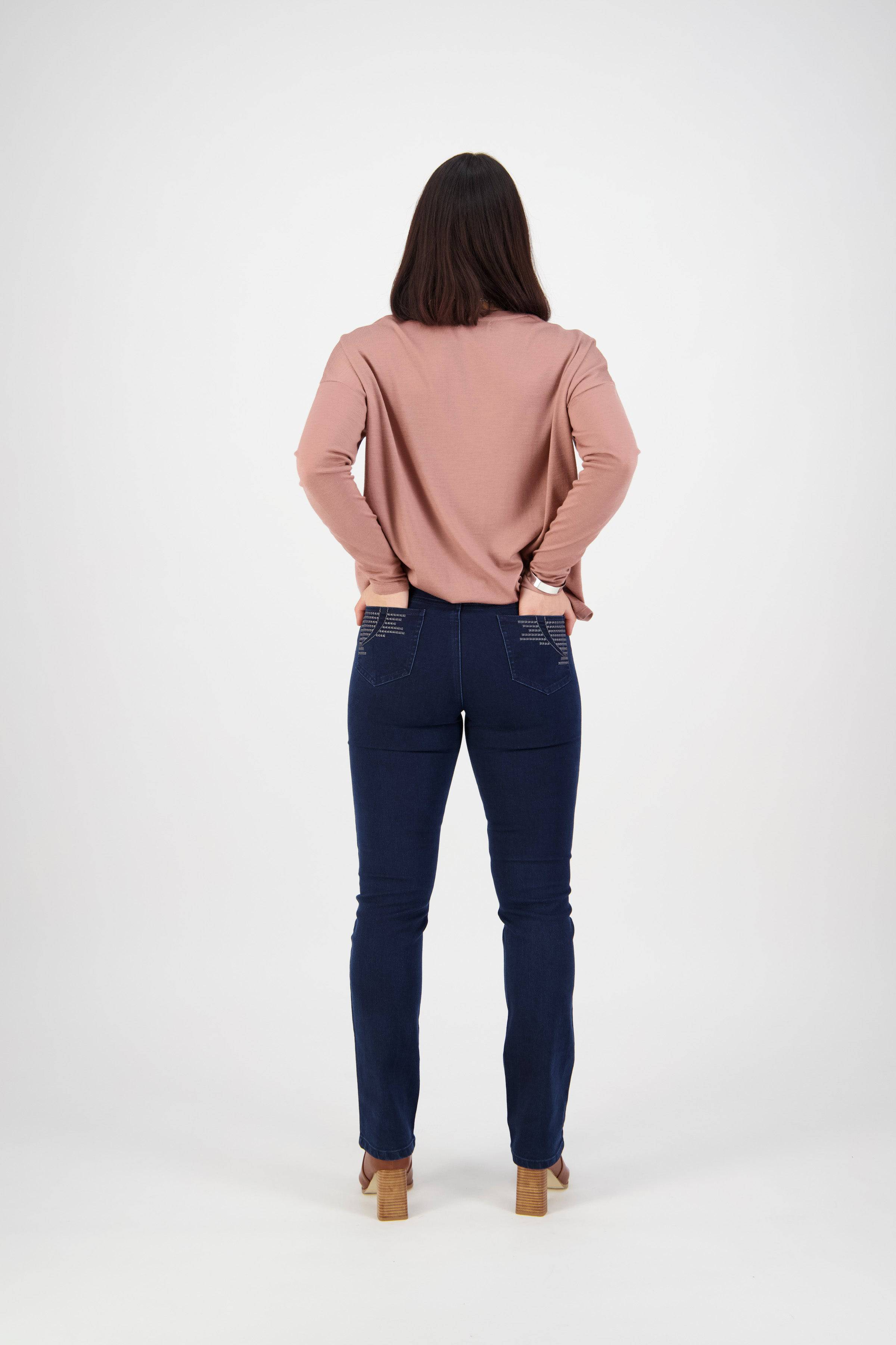 A woman with straight dark hair stands facing away, hands on hips. She wears a long-sleeved pink top, Vassalli Urban Jean - Mid Blue, and tan heeled shoes against a plain white background.