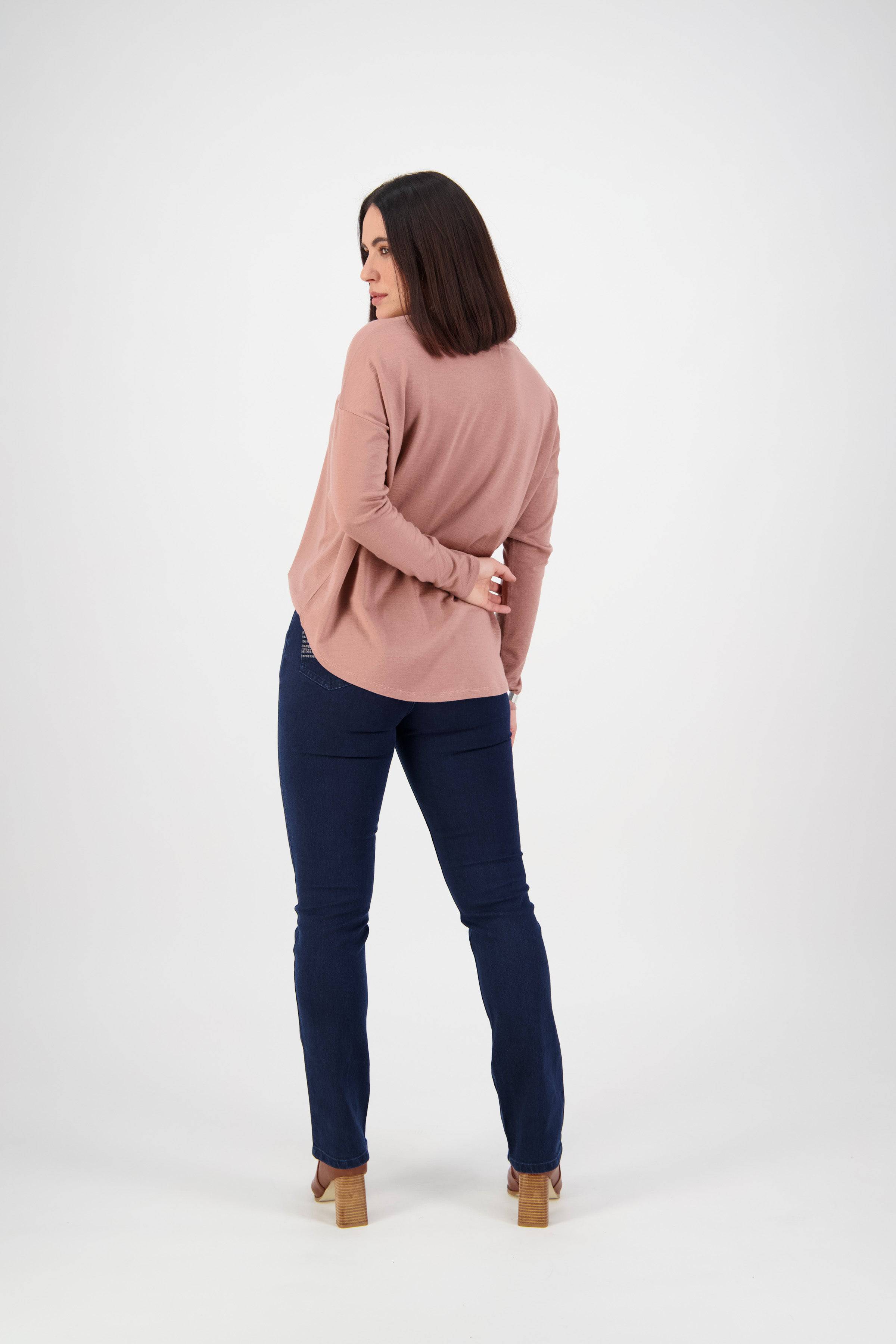A woman with straight dark hair, in a pink long-sleeve top, Vassalli Urban Jean - Mid Blue, and heeled shoes stands facing away and looks over her shoulder against a plain white background.