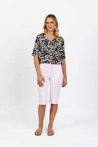 A woman with long, wavy blonde hair stands against a white background, smiling slightly. She wears a black and white patterned blouse, tan sandals, and Vassalli's Drew 3/4 Pant in white—knee-length stretch cotton shorts with an elastic waistband.