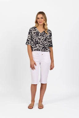 A woman with long, wavy blonde hair stands against a white background, smiling slightly. She wears a black and white patterned blouse, tan sandals, and Vassalli's Drew 3/4 Pant in white—knee-length stretch cotton shorts with an elastic waistband.