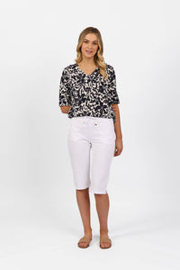 A woman with wavy blonde hair smiles gently at the camera, wearing Vassalli's Drew 3/4 Pant in white and a black-and-white patterned V-neck top, paired with brown sandals, against a plain white background.