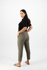 A woman with long dark hair stands in a three-quarter pose, wearing the Vassalli Denver 7/8 Pant in Army and a black top, paired with flat sandals, against a plain white background.