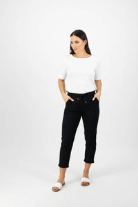 A woman stands against a white background, wearing the Vassalli Denver 7/8 Pant in black with an elastic waistband and pockets, paired with a white short-sleeve top and white slide sandals. She looks to her left with a neutral expression.