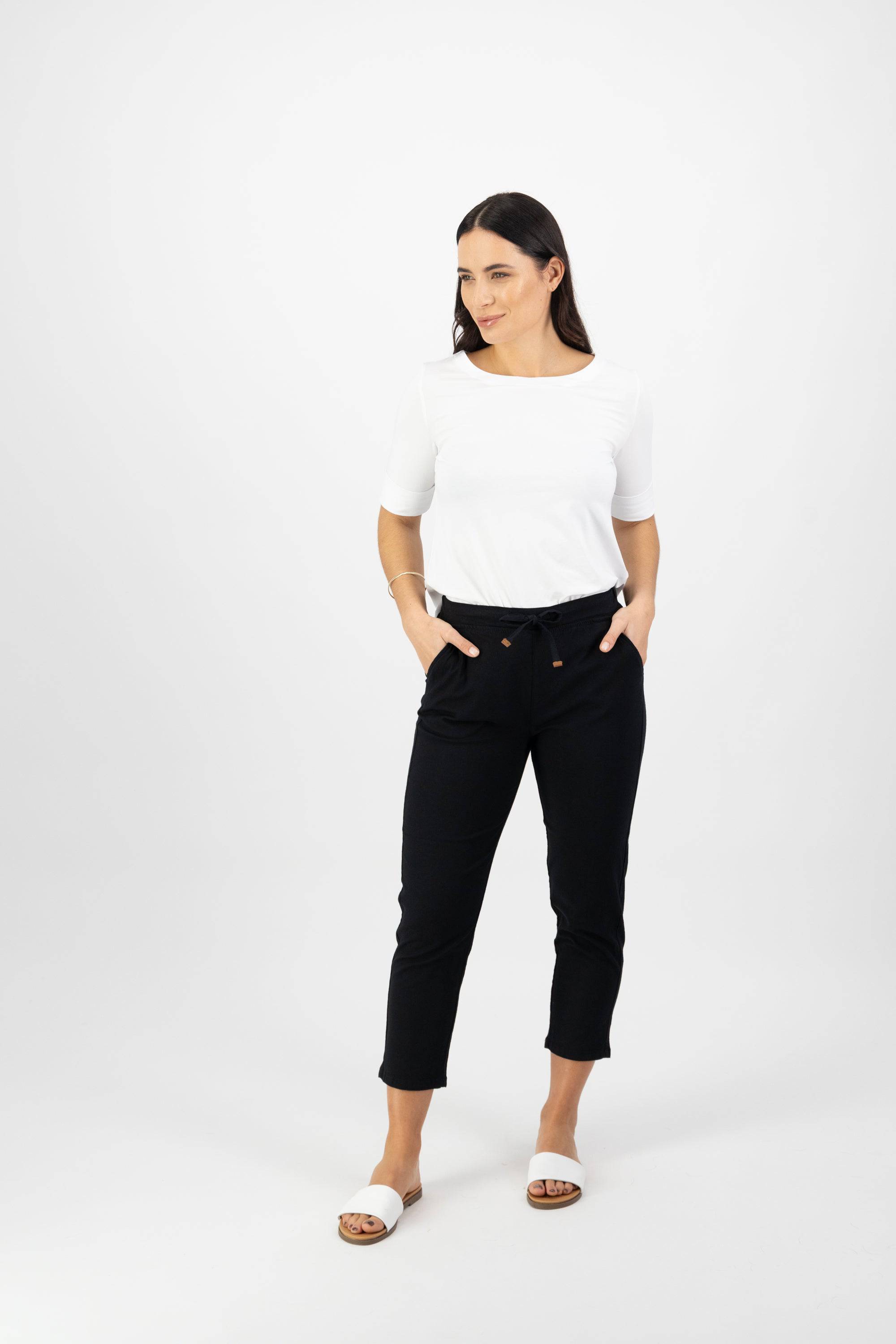 A woman stands against a white background, wearing the Vassalli Denver 7/8 Pant in black with an elastic waistband and pockets, paired with a white short-sleeve top and white slide sandals. She looks to her left with a neutral expression.