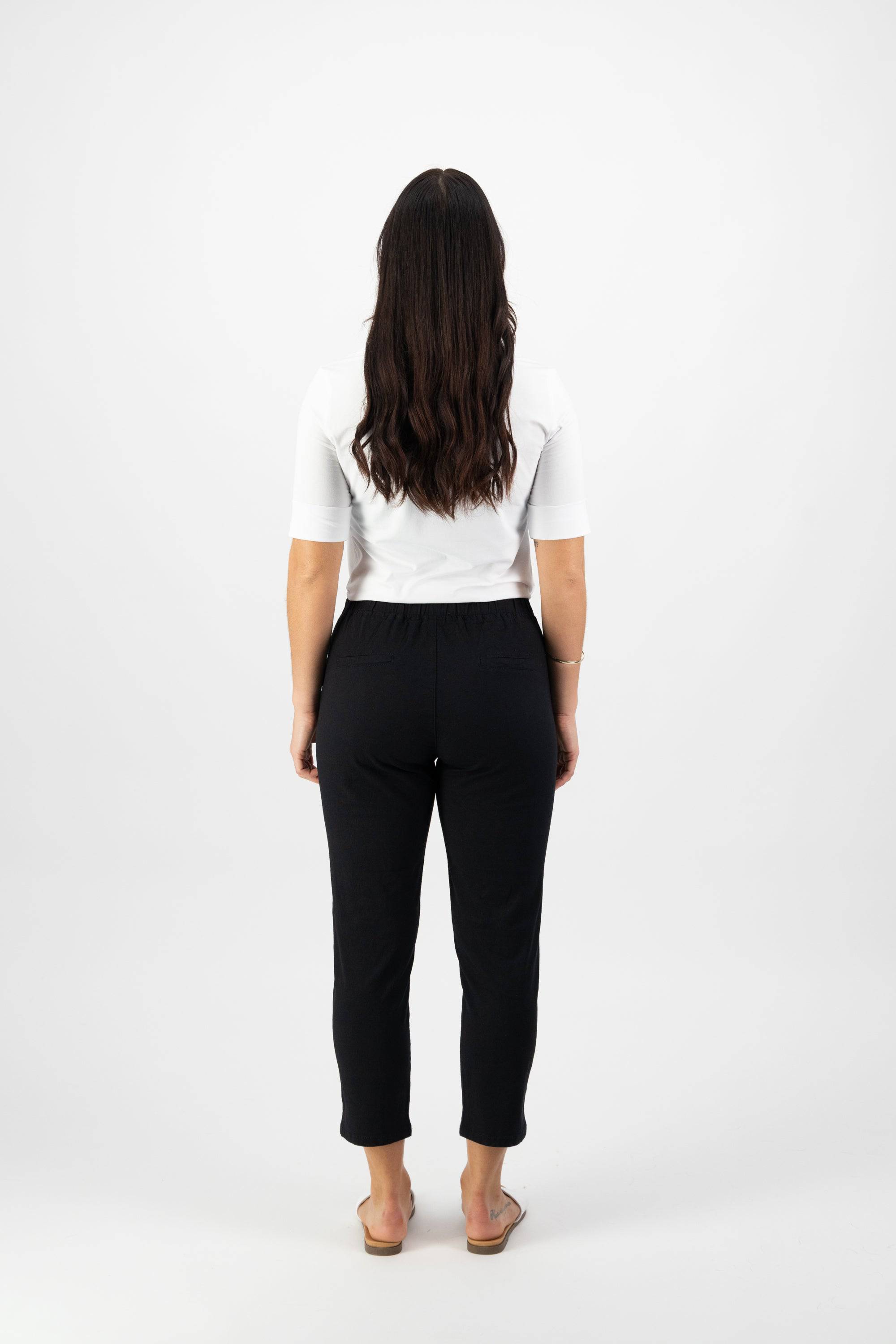 A woman with long dark hair, facing away, wears a white short-sleeve shirt and Vassalli's Denver 7/8 Pant in Black with an elastic waistband and sandals, set against a plain white background.