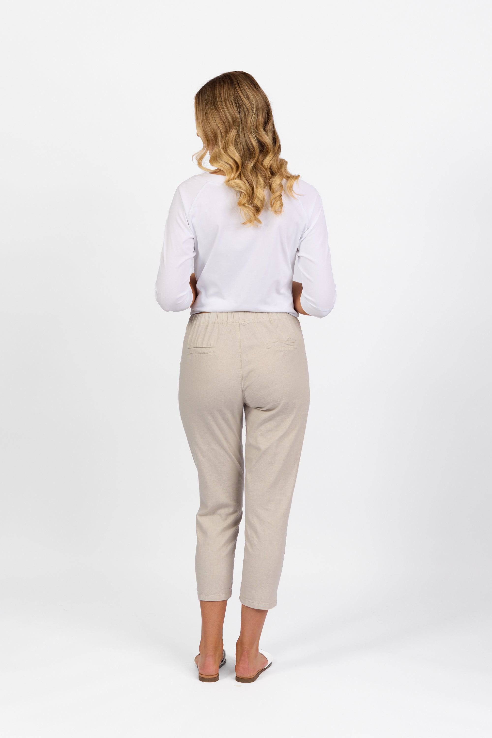 A woman with wavy blonde hair faces away, wearing a long-sleeve white top and Vassalli Denver 7/8 Pant in Stone, styled with beige sandals against a plain white background.