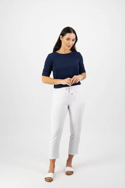 A woman with long dark hair stands against a white background, smiling slightly as she touches the waistband of her Vassalli Denver 7/8 Pant in White, styled with a navy top.