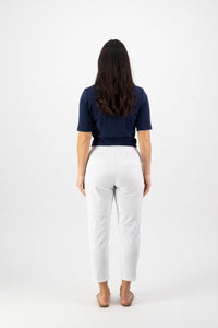 A person with long dark hair faces away, wearing a navy blue short-sleeve top, Vassalli Denver 7/8 Pant in white (cotton blend, elastic waistband), and light-colored sandals against a plain white background.