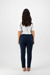A person with long dark hair stands facing away, wearing a white short-sleeve top, Vassalli Leonie 7/8 Pant in Ink, and sandals against a plain white background.