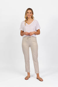 A woman smiles against a plain white background, wearing a short-sleeve white V-neck blouse, brown sandals, and Vassalli’s Leonie 7/8 Pant in Stone.