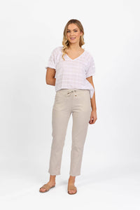 A woman stands against a white background wearing a white short-sleeve V-neck top and Vassalli Leonie 7/8 Pant in Stone, featuring a drawstring waist and slim leg stretch fit. She pairs them with tan slide sandals and has one arm behind her back.