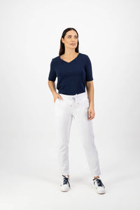 A woman stands against a white background, wearing the Vassalli Leonie 7/8 Pant in White with a navy blue short-sleeve top and black-and-white sneakers, looking slightly to the side with a neutral expression.