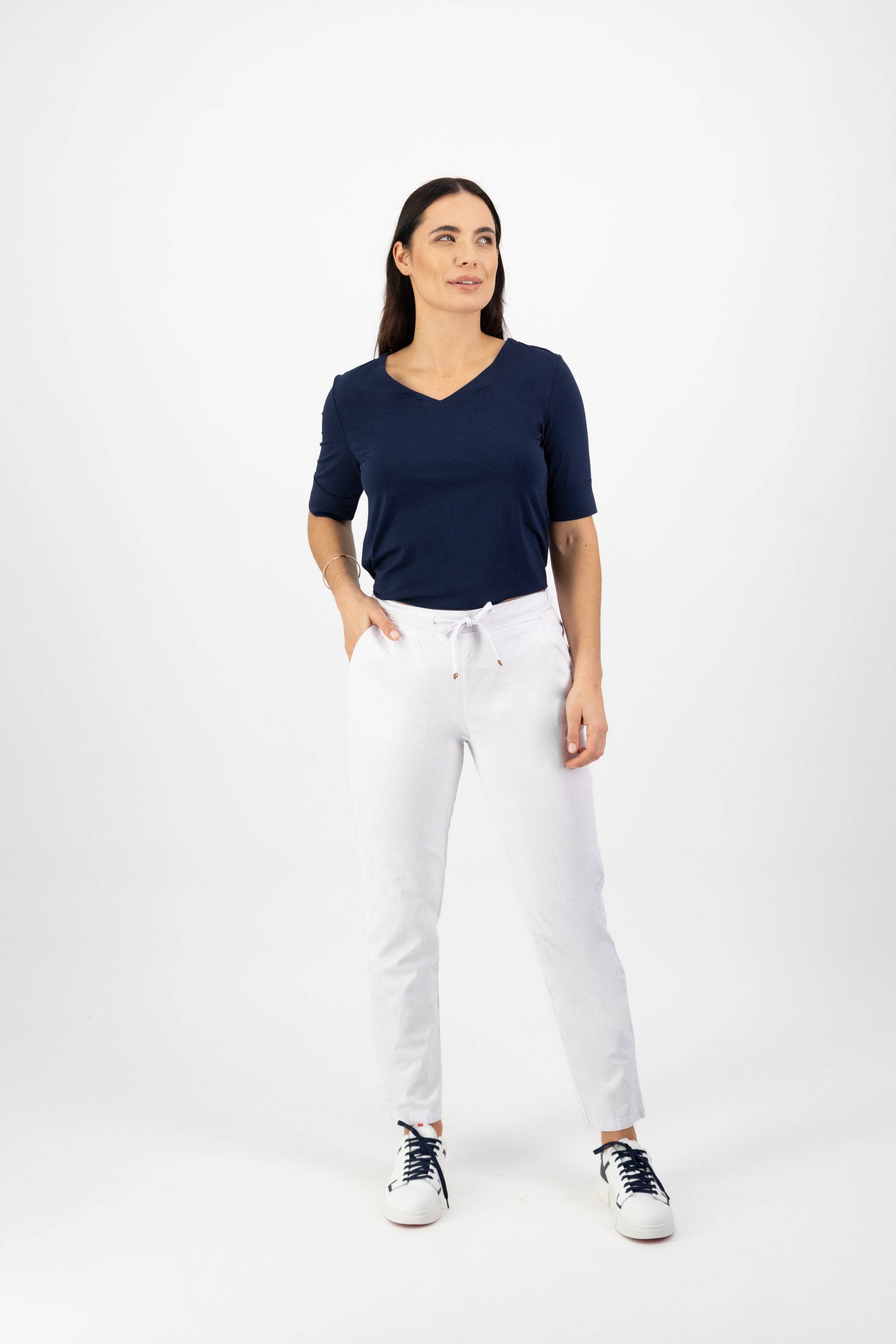 A woman stands against a white background, wearing the Vassalli Leonie 7/8 Pant in White with a navy blue short-sleeve top and black-and-white sneakers, looking slightly to the side with a neutral expression.