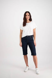 A woman stands against a white background, wearing the Vassalli Alfa Short in indigo denim with contrast stitch—a summer staple—paired with a white t-shirt, white sneakers, and her long brown hair worn loosely.