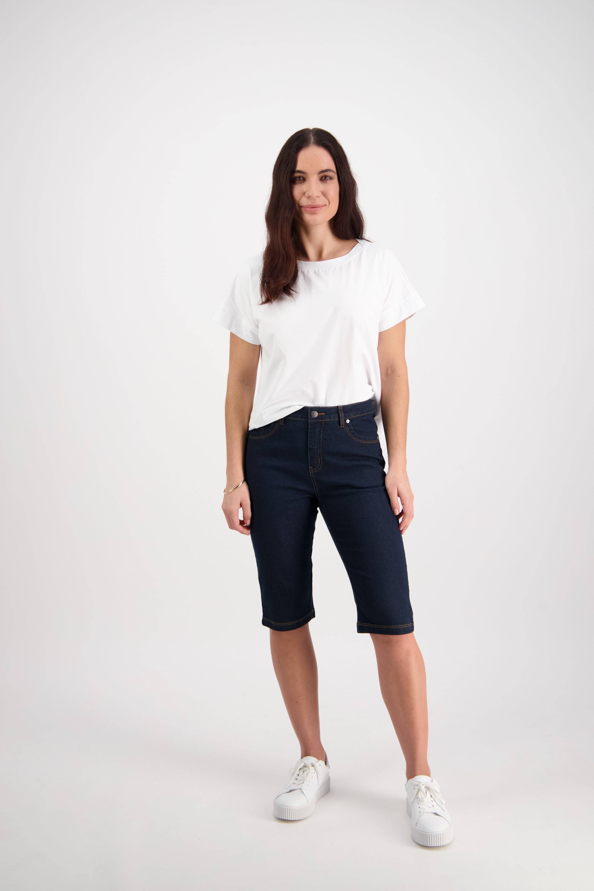 A woman stands against a white background, wearing the Vassalli Alfa Short in indigo denim with contrast stitch—a summer staple—paired with a white t-shirt, white sneakers, and her long brown hair worn loosely.