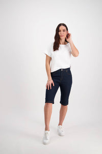 A woman with long brown hair stands against a white background wearing Vassalli's Alfa Short in Indigo Denim (Contrast Stitch), a white short-sleeve tee, and white sneakers, touching her ear with one hand.