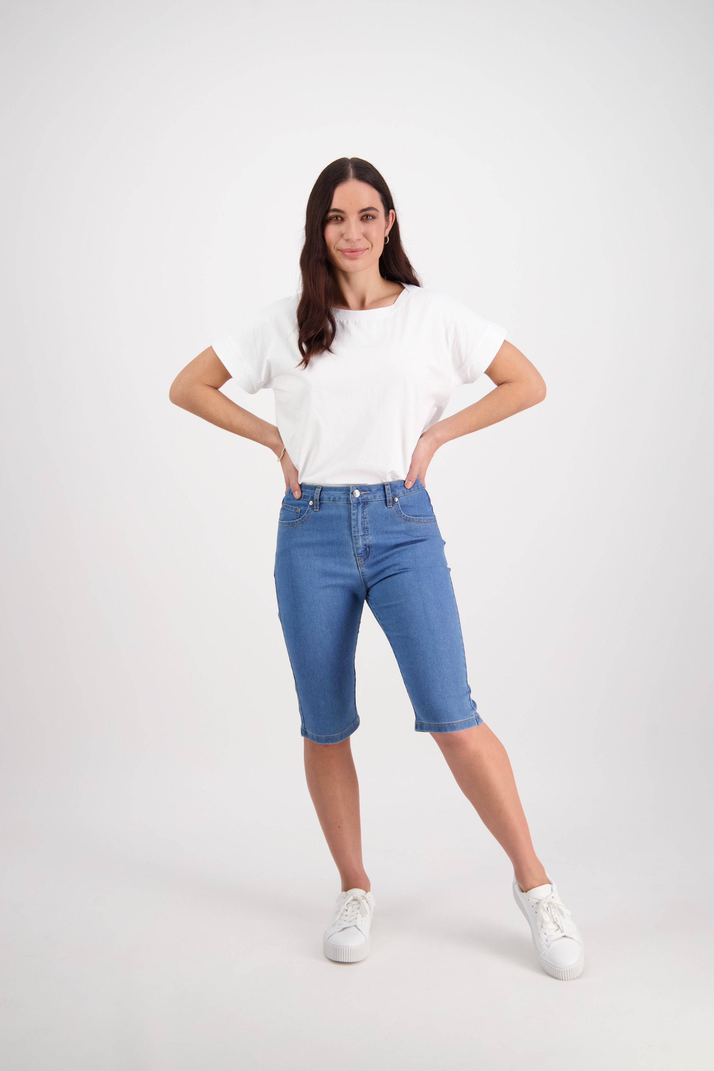 A woman poses against a white background wearing a white t-shirt, Vassalli's Alfa Short - Light Denim (Contrast Stitch), and white sneakers. She smiles confidently, hands on hips—a fresh summer staple look.