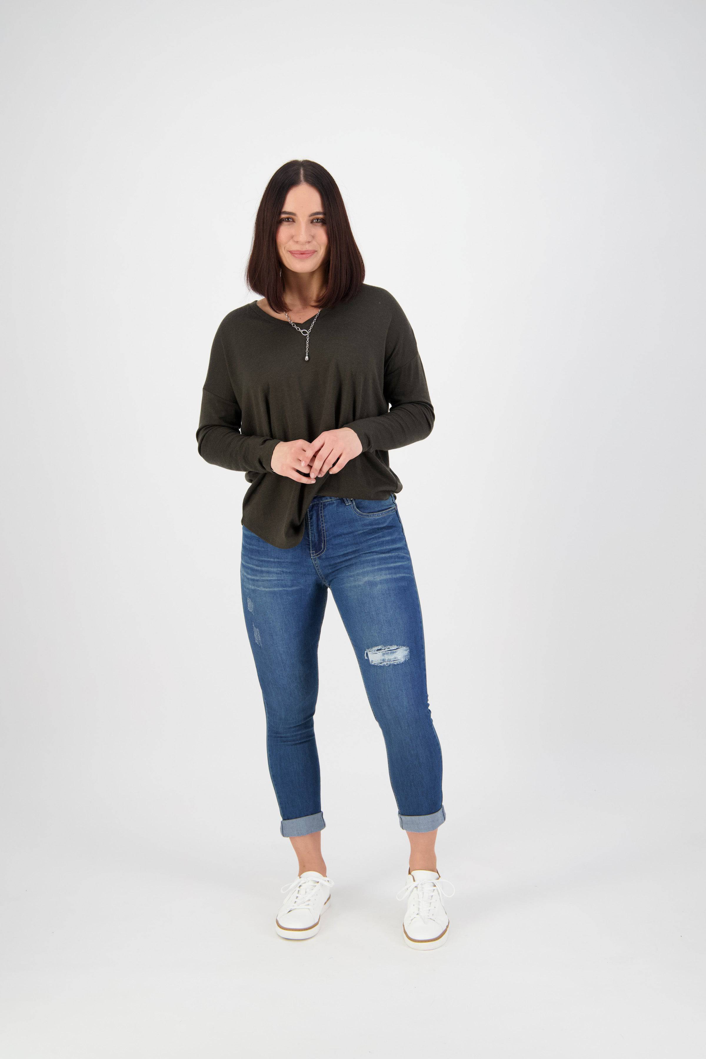 A woman with shoulder-length dark hair stands against a plain white background, smiling softly. She wears a dark long-sleeve shirt, Vassalli Coast Jean - Blue Denim with cuffed ankles, and white sneakers, her hands together.