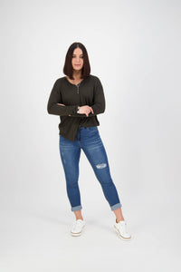 A woman with straight, shoulder-length dark hair stands confidently with arms crossed, wearing a dark long-sleeve top, Vassalli Coast Jean - Blue Denim, and white sneakers against a plain white background.