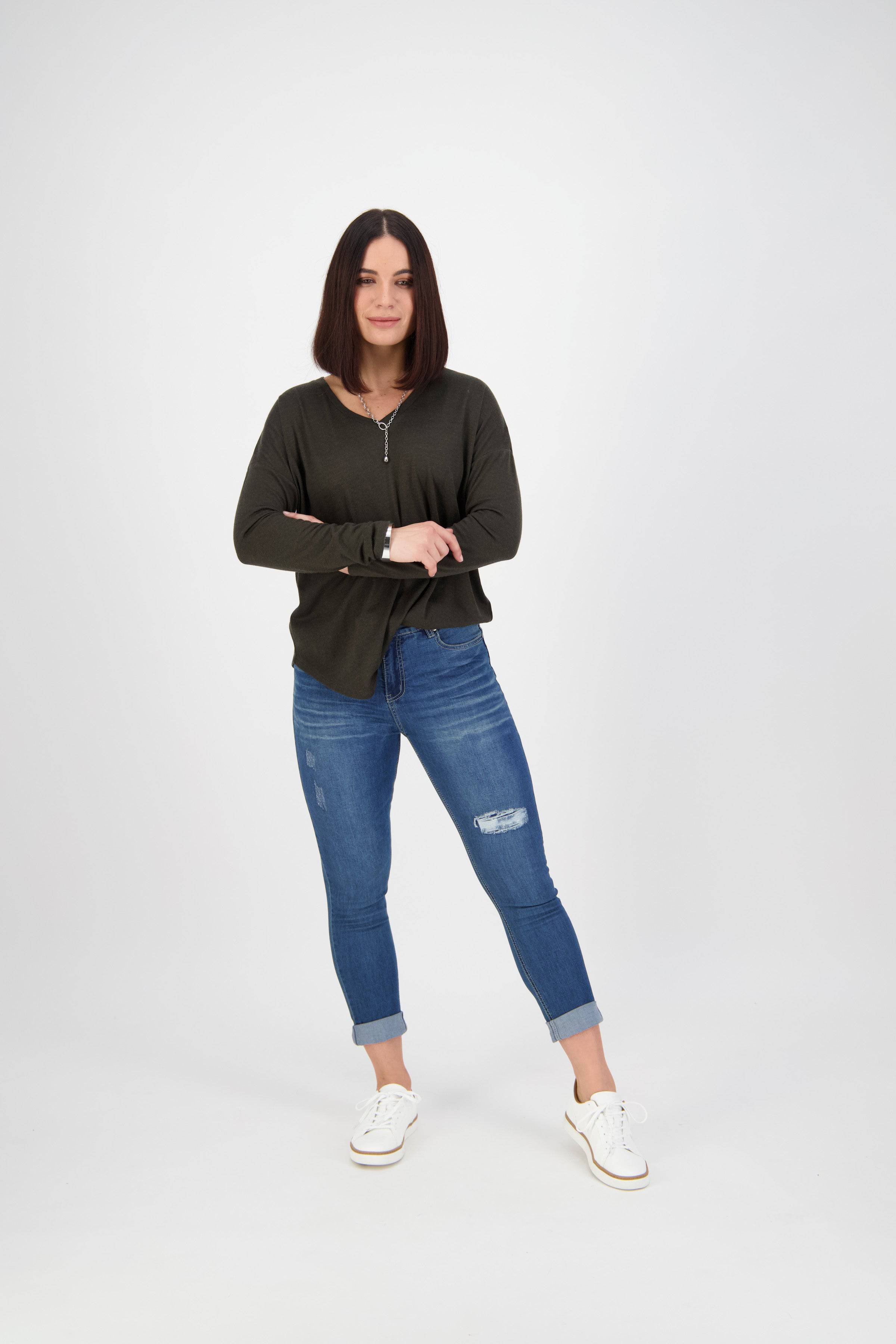 A woman with straight, shoulder-length dark hair stands confidently with arms crossed, wearing a dark long-sleeve top, Vassalli Coast Jean - Blue Denim, and white sneakers against a plain white background.