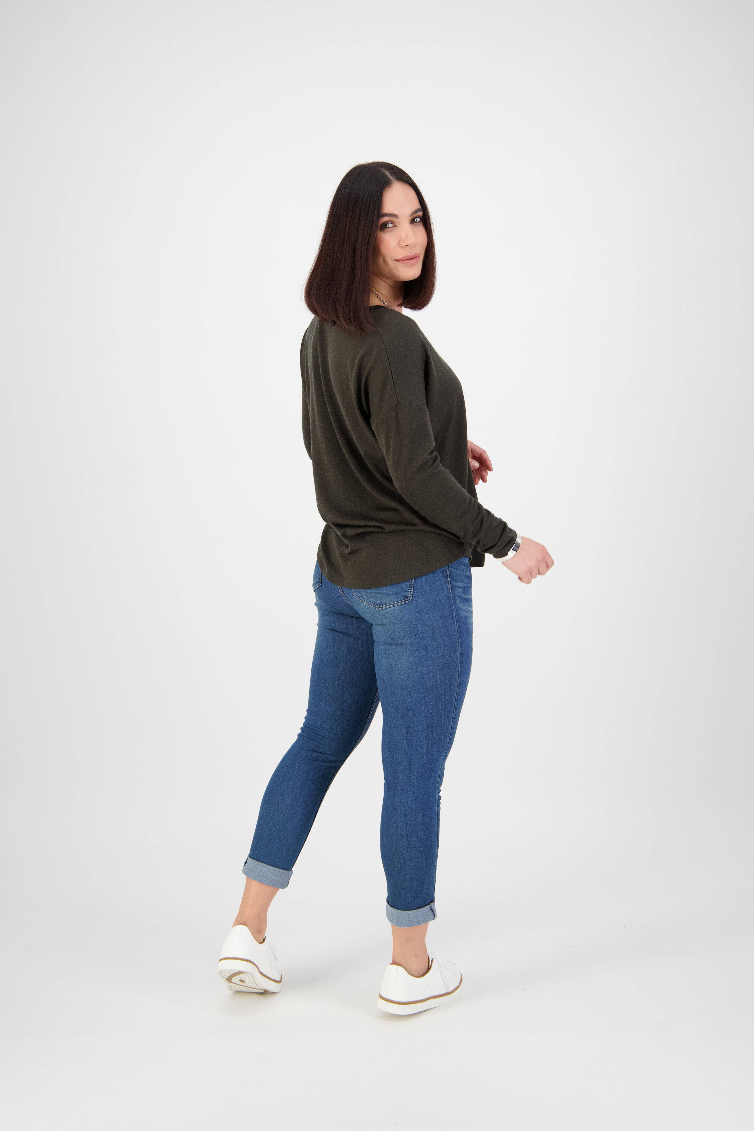 A woman with straight, shoulder-length dark hair stands slightly turned to the side, wearing a dark long-sleeve top, Vassalli Coast Jean in blue denim, and white sneakers, posing against a plain white background.