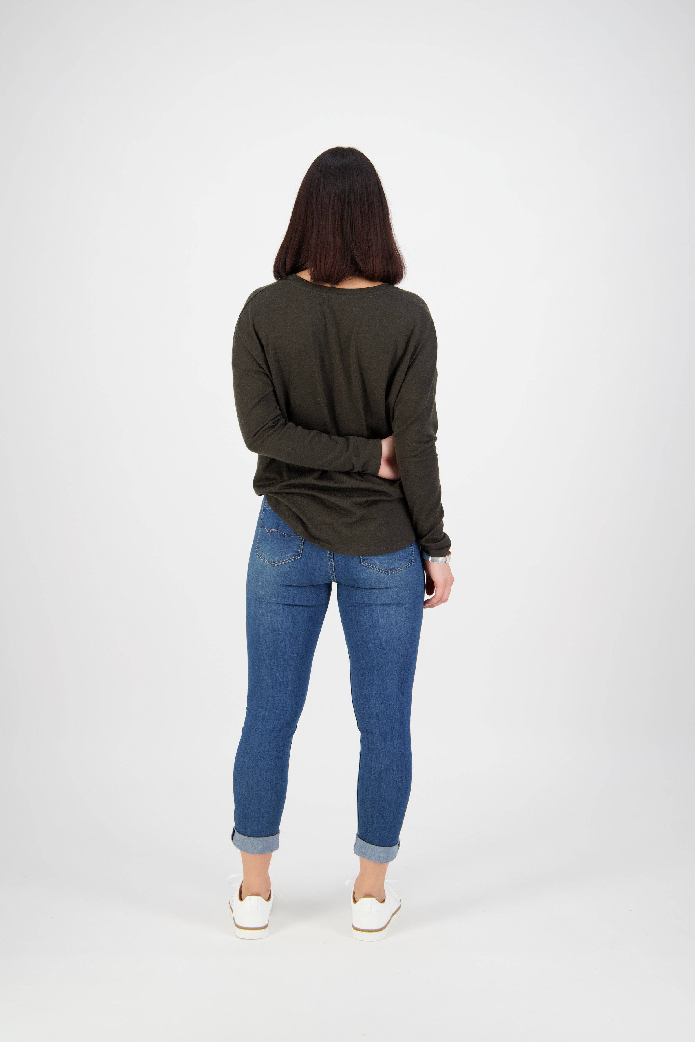 A person with straight dark hair, wearing a dark long-sleeve shirt, white shoes, and Vassalli Coast Jean - Blue Denim, stands facing away from the camera with one hand behind their back. The background is plain white.