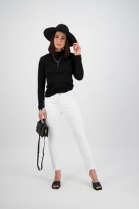 A woman poses against a plain background wearing a black wide-brimmed hat, black long-sleeve top, Vassalli Tide Jean in white denim, black heeled sandals, and holding a small black handbag.