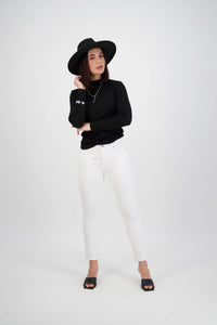 A woman poses against a white background, wearing a black wide-brim hat, black long-sleeve top, and Vassalli Tide Jean in White Denim with black open-toe shoes. She stands with one hand on her hat and her other arm crossed.