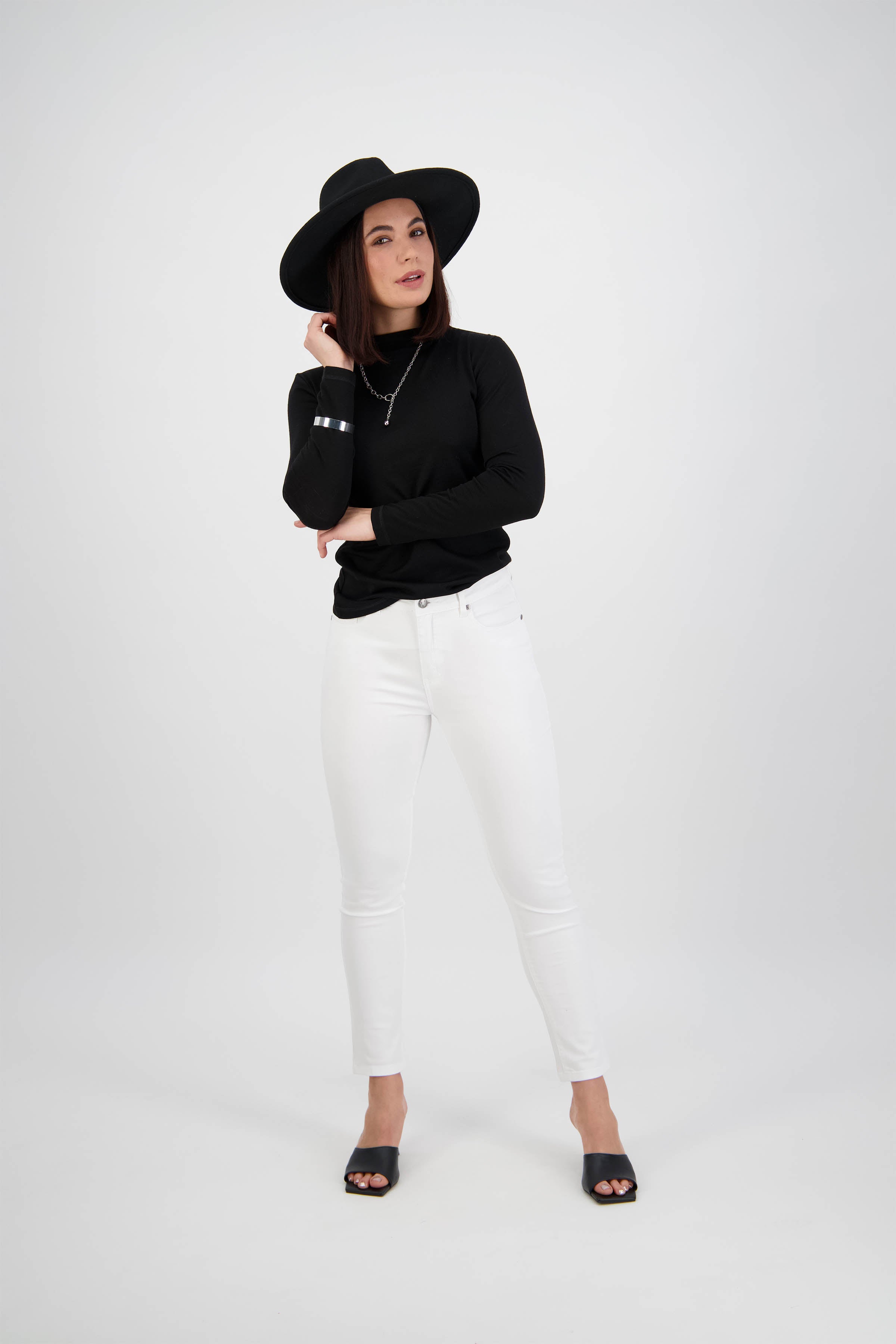 A woman poses against a white background, wearing a black wide-brim hat, black long-sleeve top, and Vassalli Tide Jean in White Denim with black open-toe shoes. She stands with one hand on her hat and her other arm crossed.