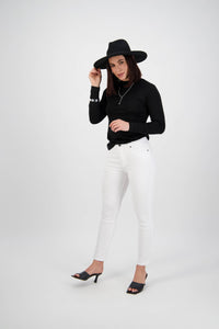 A woman wearing a black wide-brim hat, black long-sleeve top, Vassalli Tide Jean in white denim, and black heeled sandals poses confidently against a plain white background.