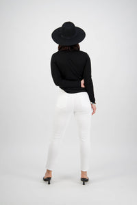 A person stands with their back to the camera, wearing a black wide-brimmed hat, black long-sleeve top, Vassalli Tide Jean in white denim, and black heels against a plain white background.