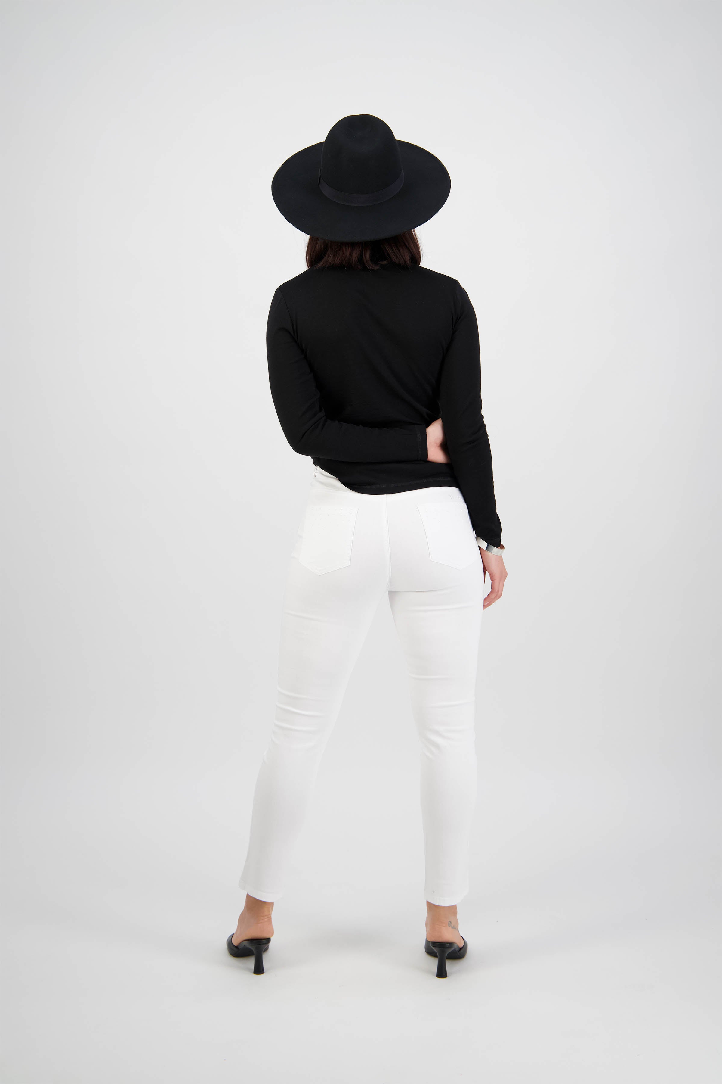 A person stands with their back to the camera, wearing a black wide-brimmed hat, black long-sleeve top, Vassalli Tide Jean in white denim, and black heels against a plain white background.