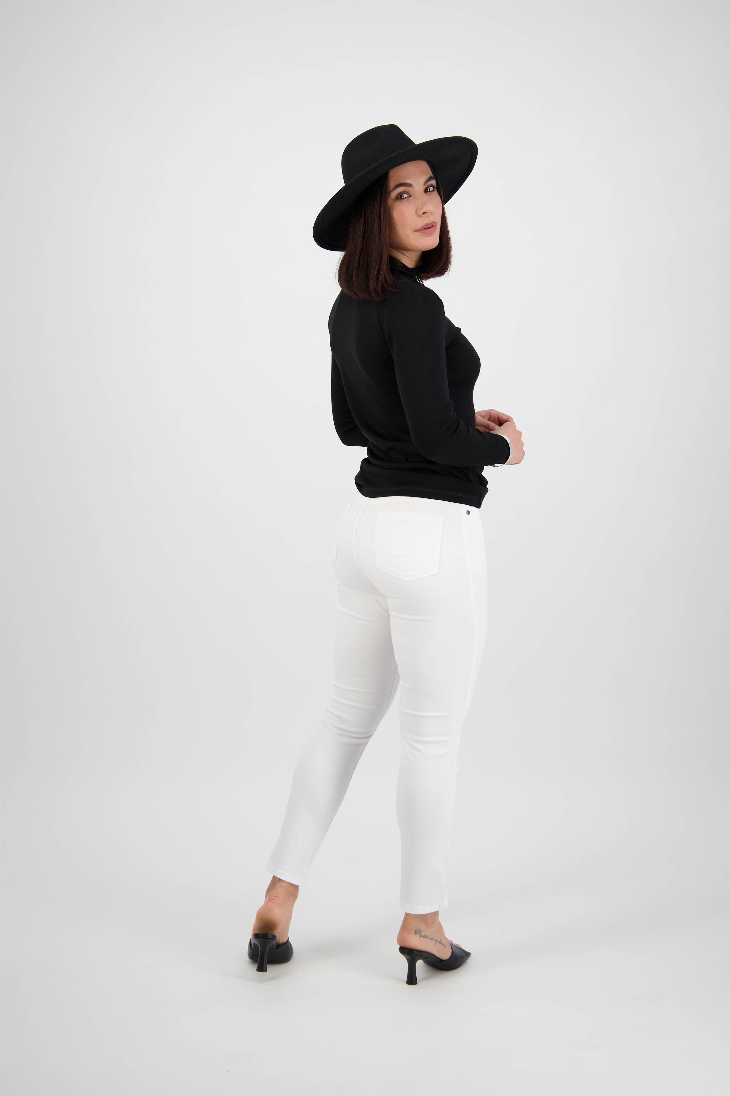 A woman poses against a white background wearing a black wide-brimmed hat, black long-sleeve top, Vassalli Tide Jean in white denim, and black heels, glancing over her shoulder toward the camera.