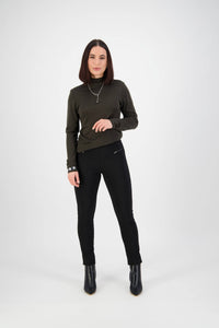 A woman with straight brown hair stands against a white background, wearing a dark green long-sleeve top, Vassalli Concept Pant - Black, black ankle boots, and silver jewelry. She faces forward with a neutral expression.
