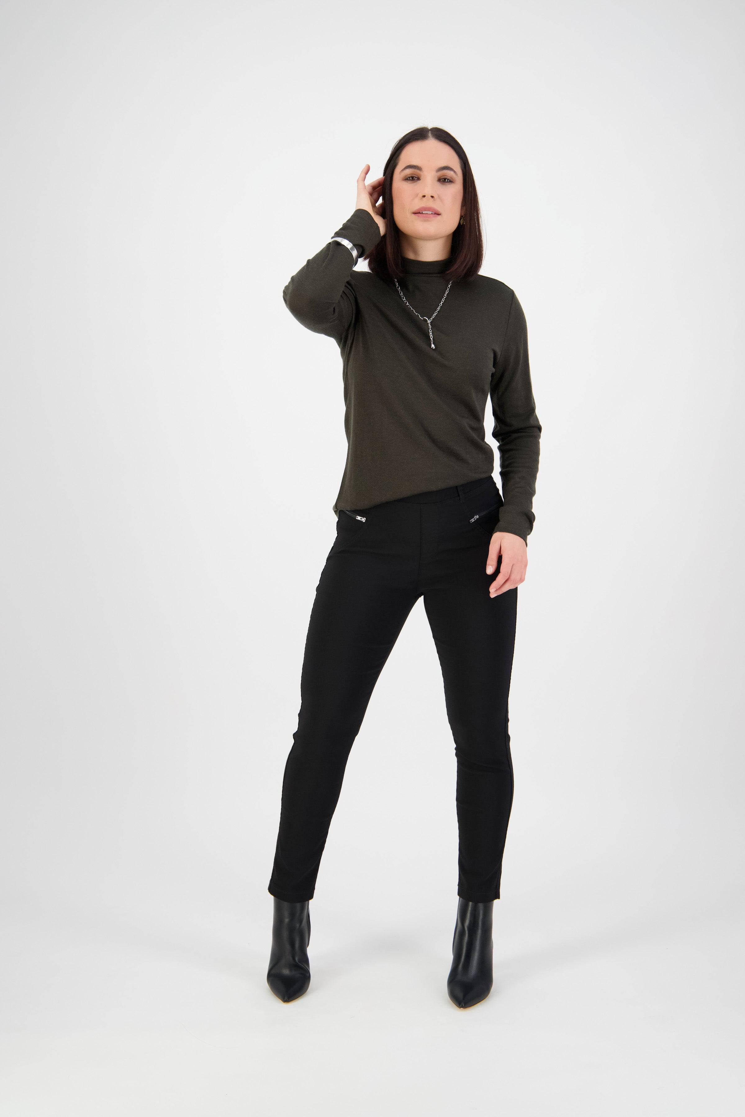 A woman with straight dark hair stands confidently before a white background wearing a dark long-sleeve top, Vassalli Concept Pant - Black, ankle boots, and a necklace, with one hand touching her hair.