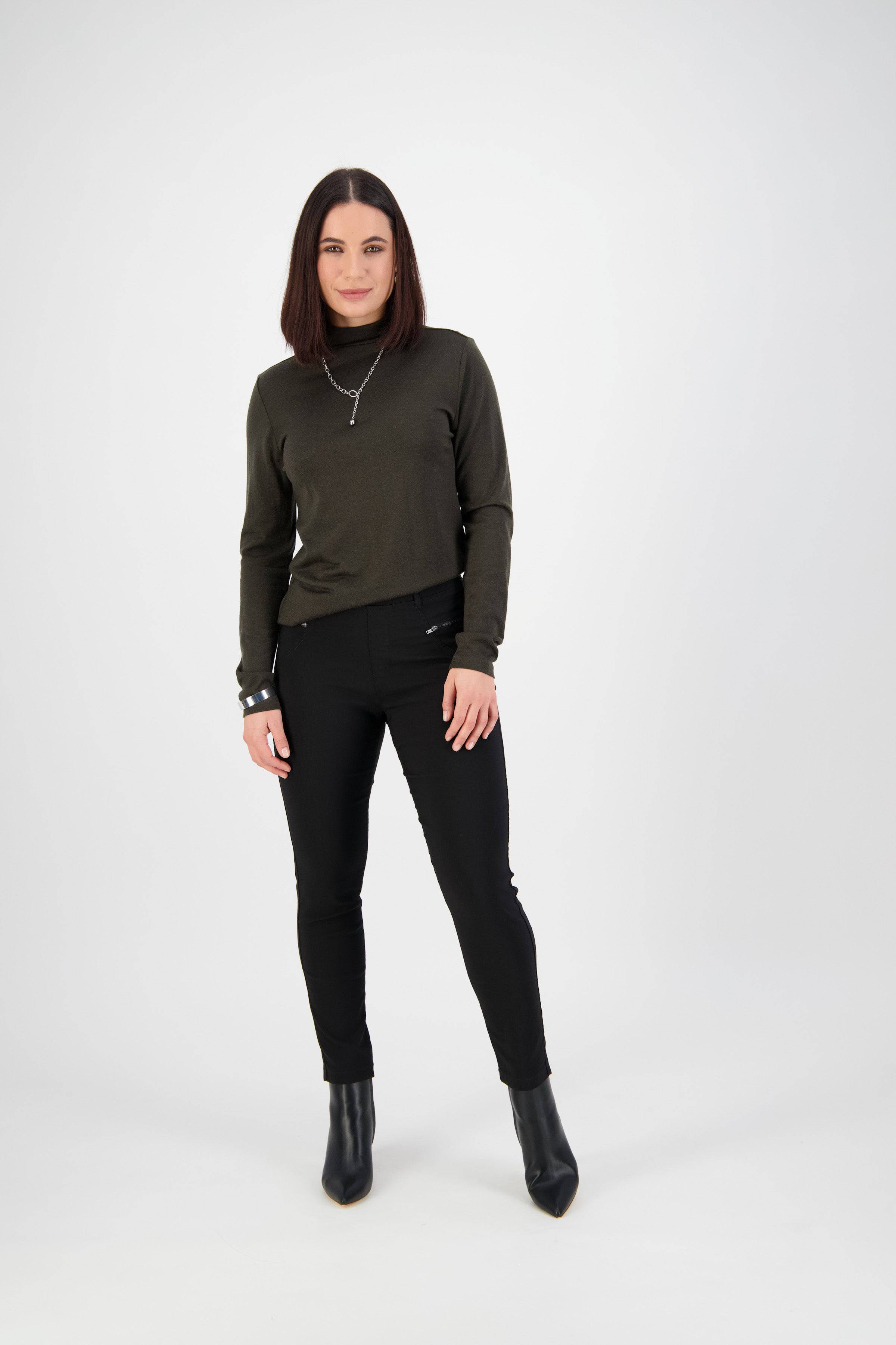 A woman with straight, shoulder-length dark hair stands against a plain white background, wearing a dark long-sleeve turtleneck top, Vassalli Concept Pant - Black, and black ankle boots. She has a neutral expression and relaxed posture.