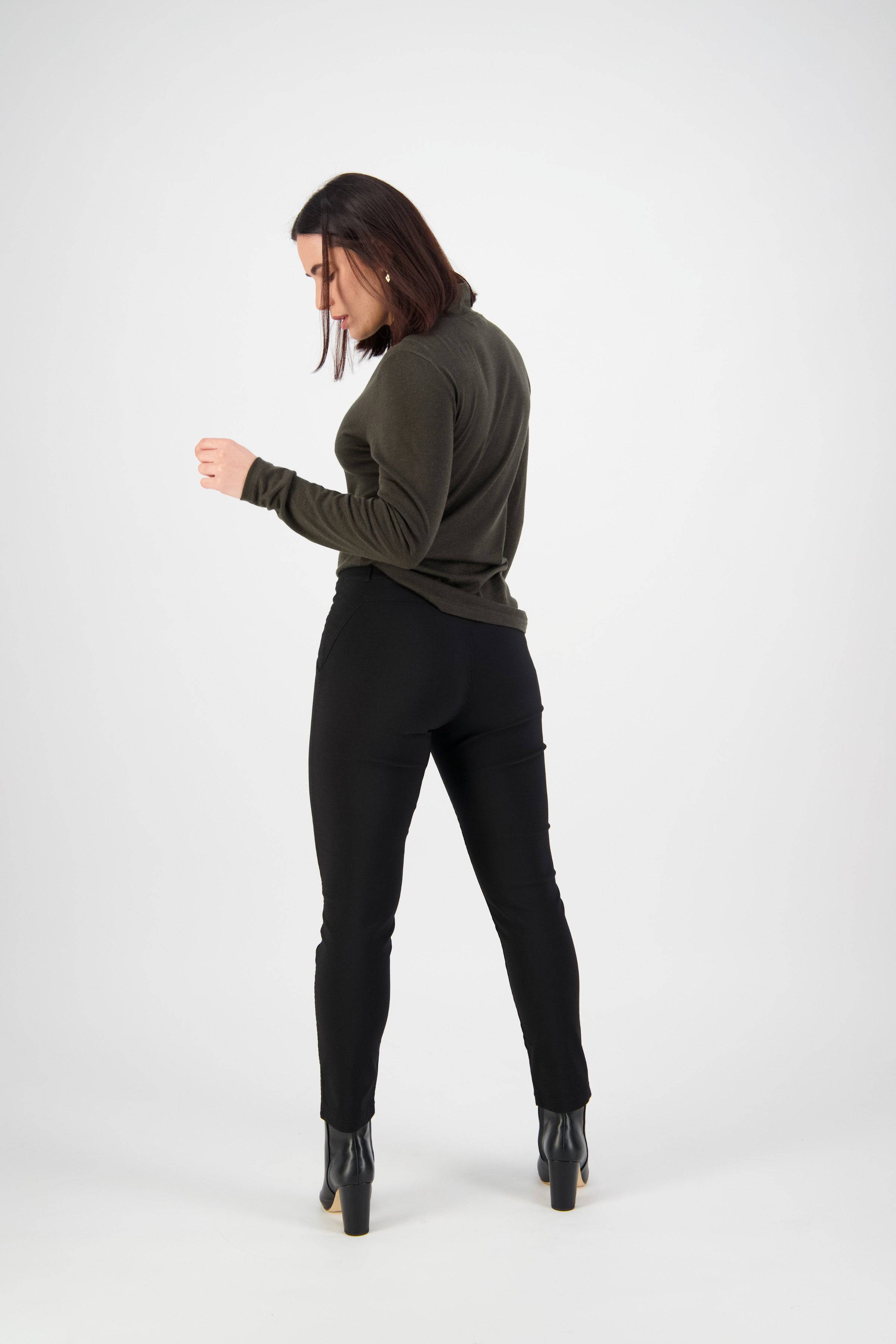 A woman with shoulder-length dark hair, in a dark green long-sleeve top, Vassalli Concept Pant - Black, and black heeled boots stands facing away and looking down against a plain white background.