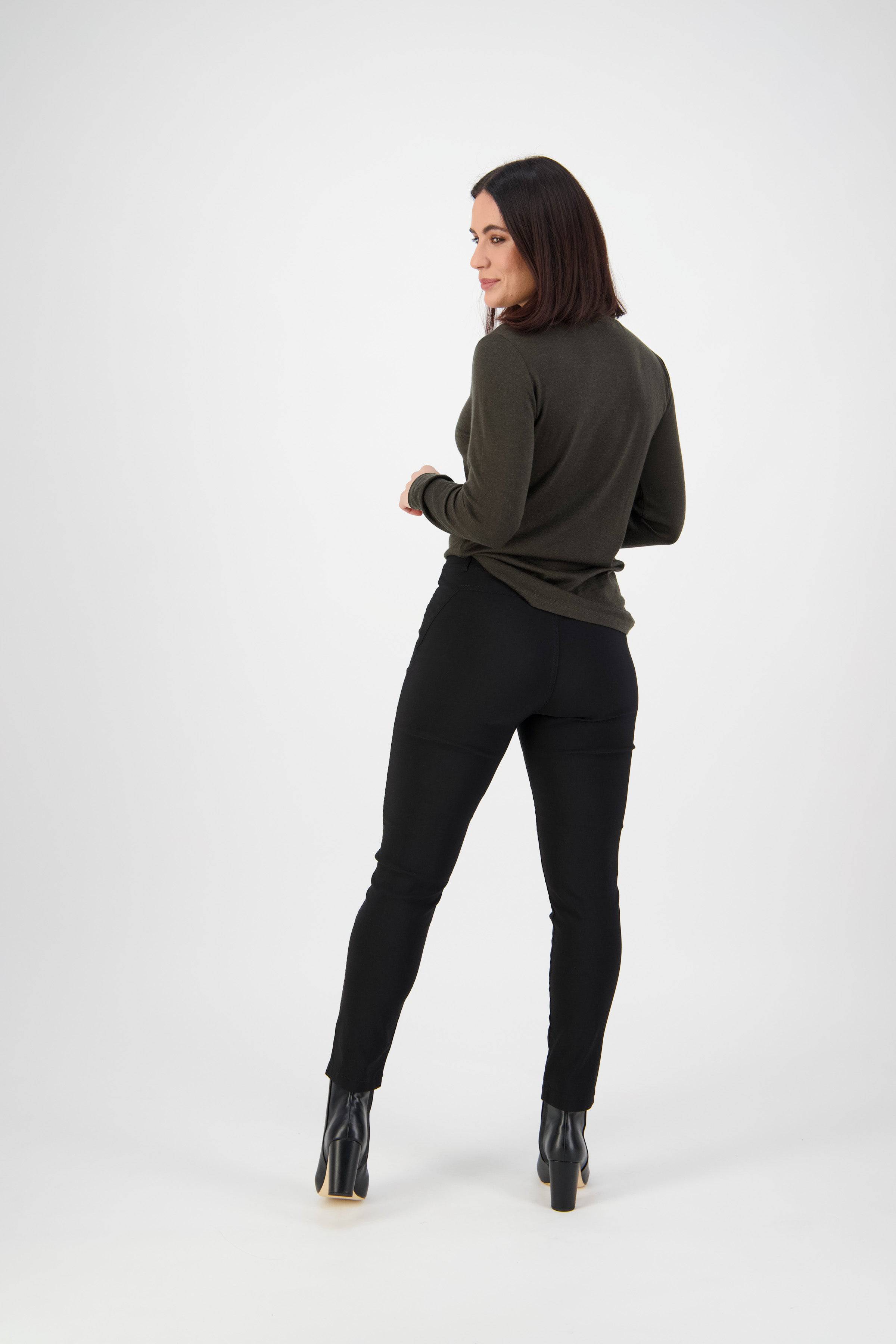 A woman with shoulder-length dark hair, wearing a dark green long-sleeve top, Vassalli Concept Pant in black, and black heeled boots stands facing away, turning her head slightly to the side against a plain white background.