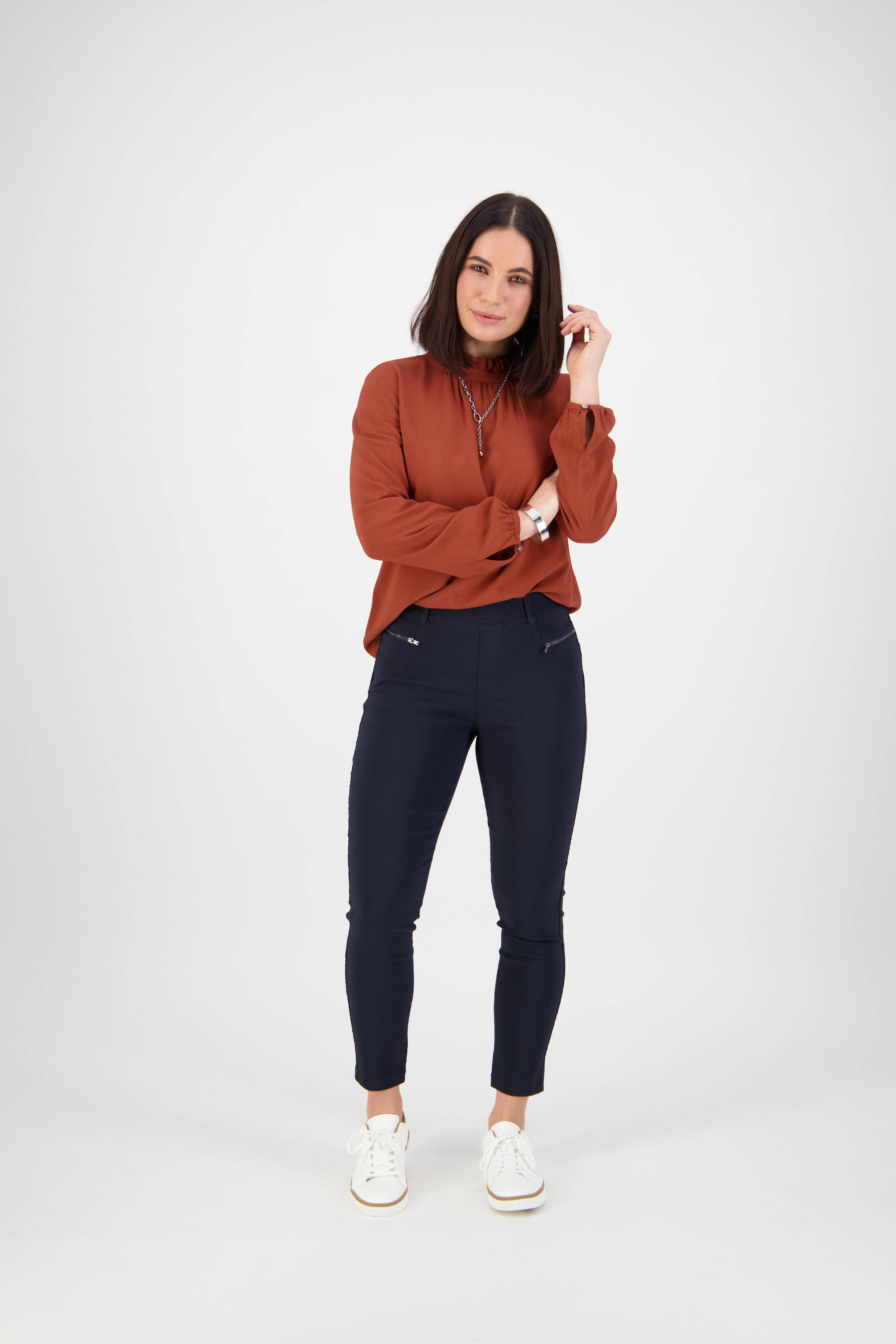 A woman with shoulder-length dark hair stands against a white background, wearing a rust-colored long-sleeve blouse, Vassalli Concept Pant in navy, and white sneakers, smiling slightly as she touches her hair.
