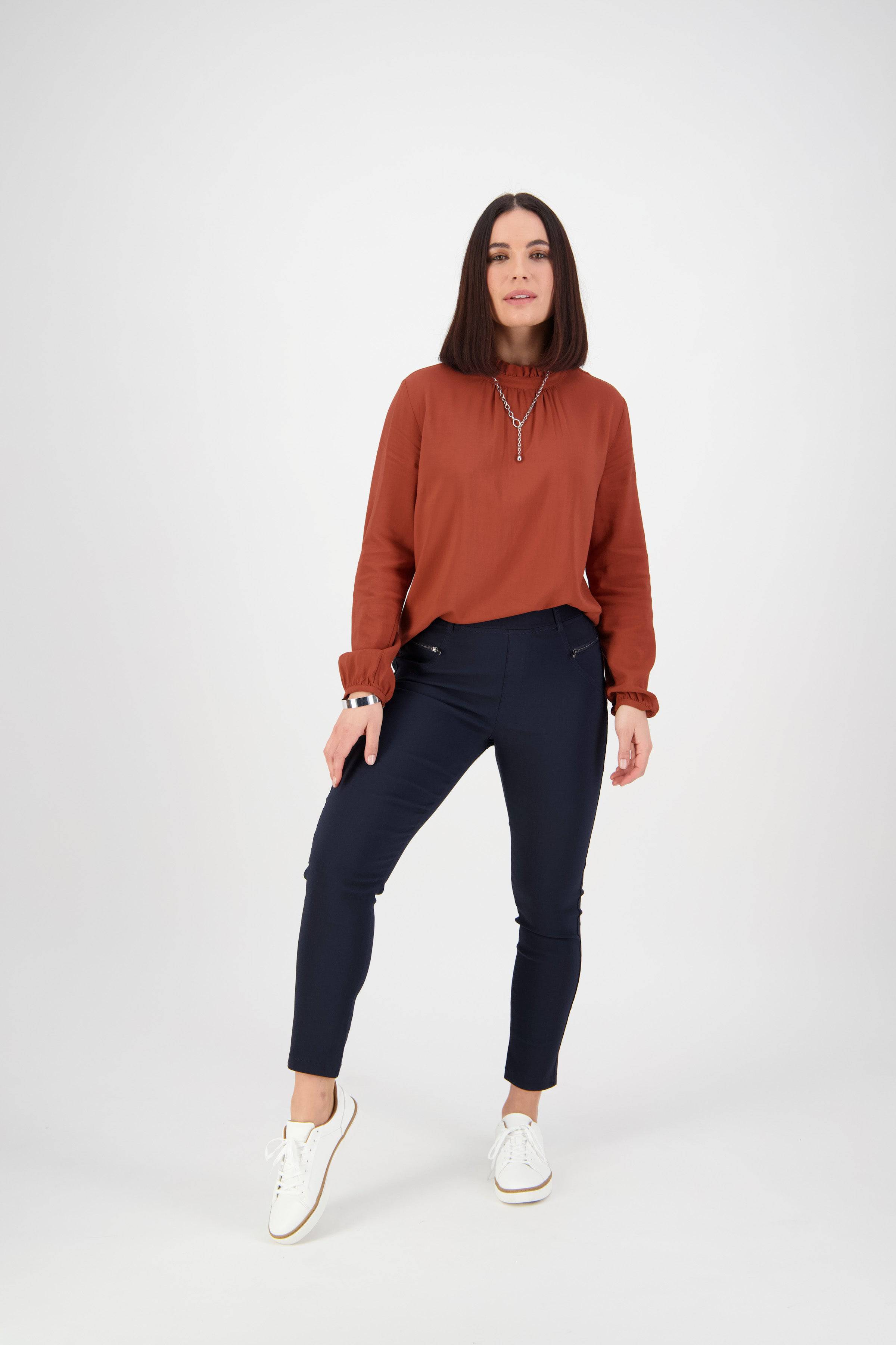 A woman with straight dark hair stands against a white background, wearing a rust long-sleeve blouse, Vassalli Concept Pant in navy, and white sneakers. She poses relaxedly with one hand on her hip and a slight smile.