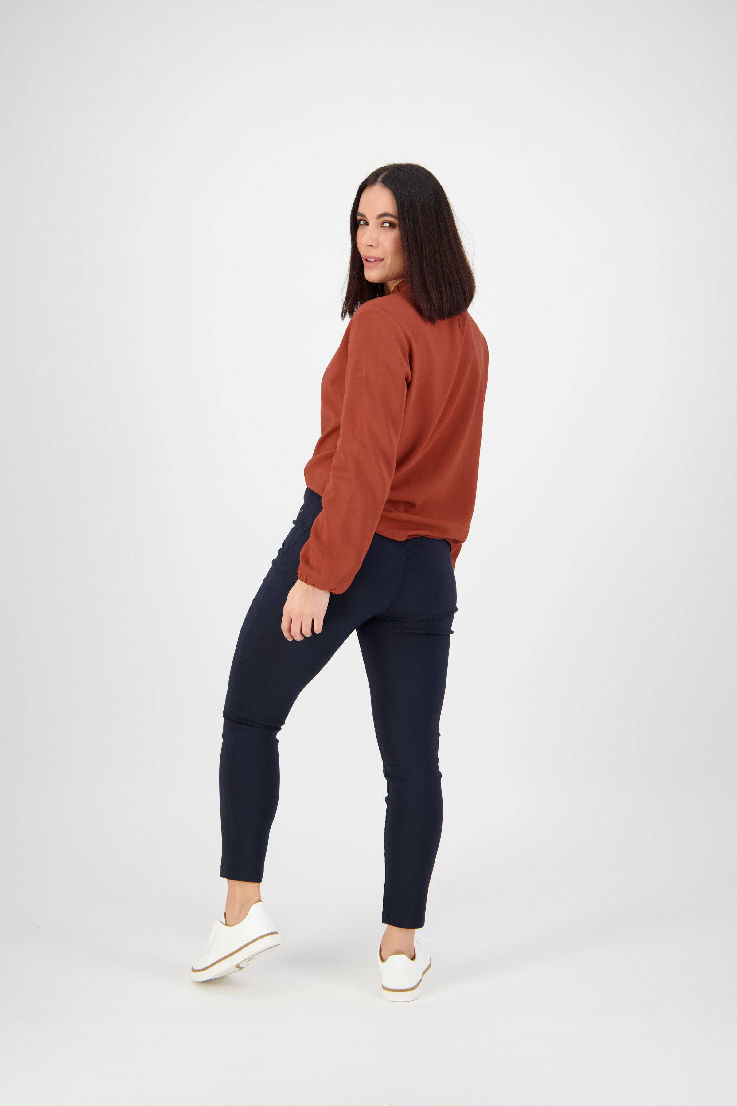 A woman with shoulder-length dark hair poses in a rust long-sleeve top, Vassalli Concept Pant in navy, and white slip-on shoes, looking over her shoulder against a plain white background.