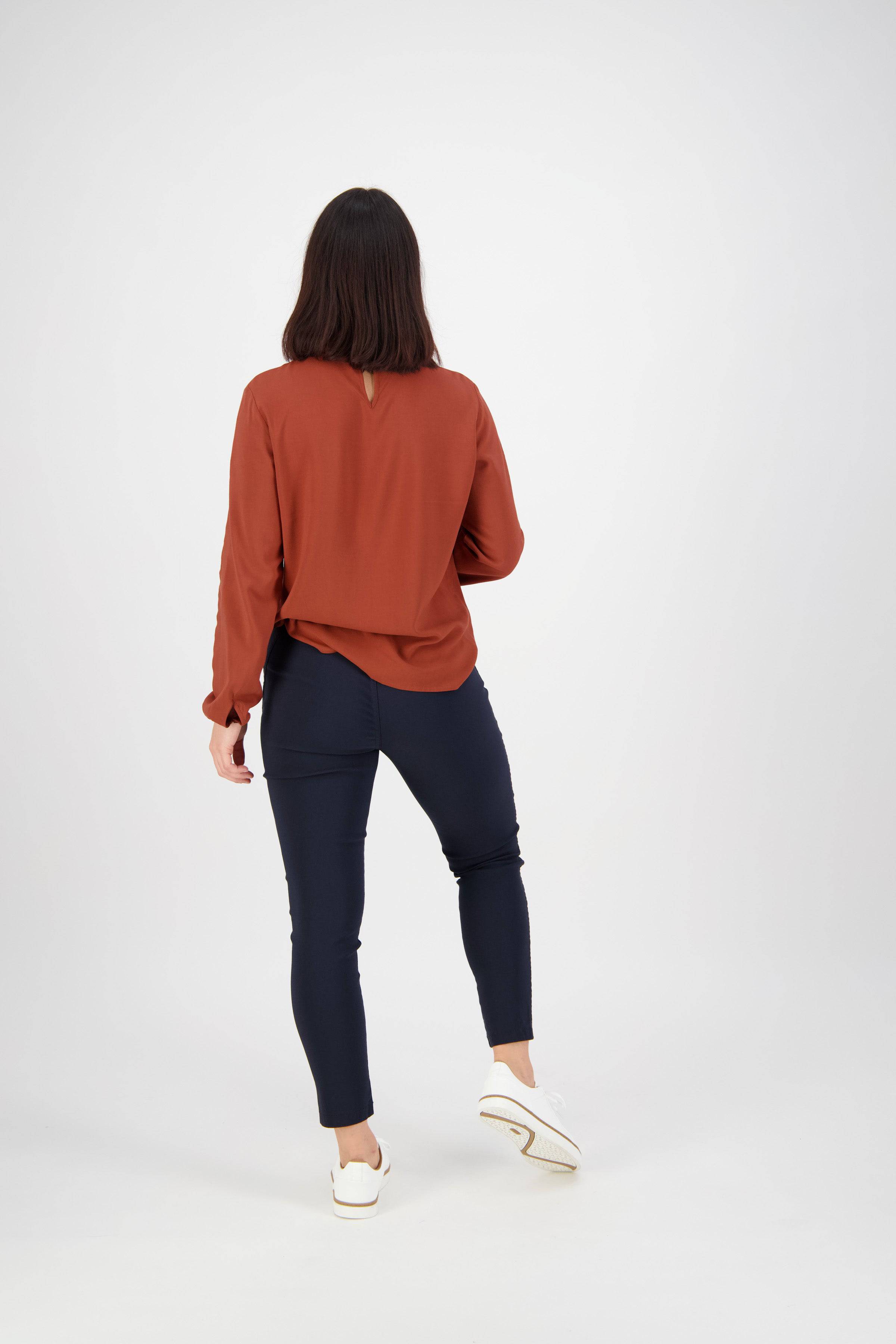 A woman with straight dark hair, facing away, wears a long-sleeved reddish-brown blouse, Vassalli Concept Pant - Navy, and white sneakers against a plain light background.