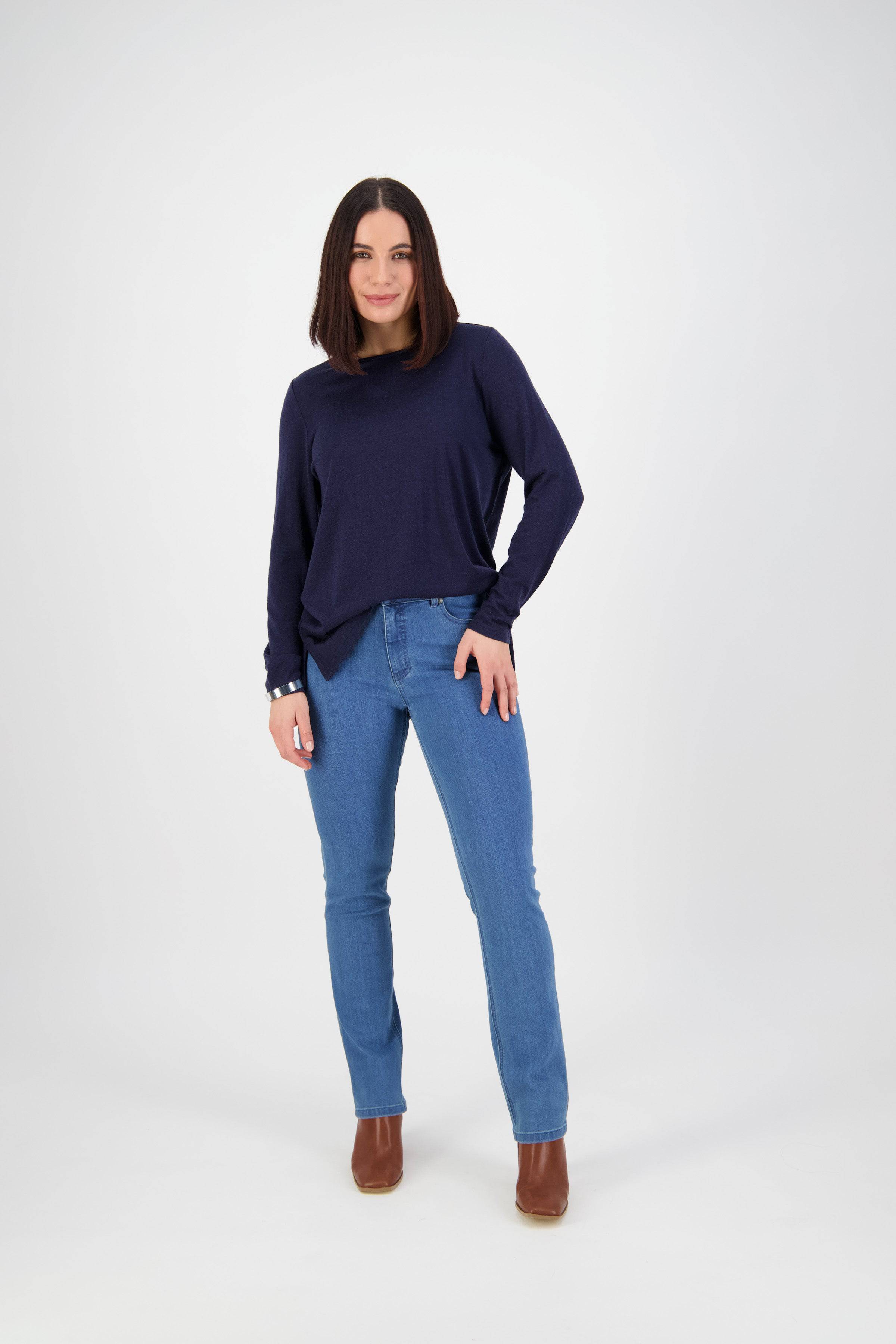 A woman with straight dark hair poses in a navy long-sleeve top, Vassalli Onset Jean - Plain New Blue slim leg jeans, and brown ankle boots against a plain white background.