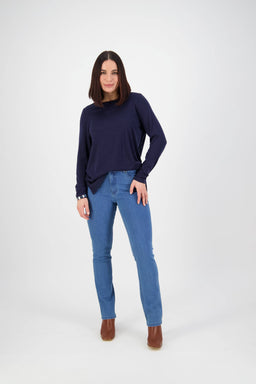 A woman with straight dark hair poses in a navy long-sleeve top, Vassalli Onset Jean - Plain New Blue slim leg jeans, and brown ankle boots against a plain white background.